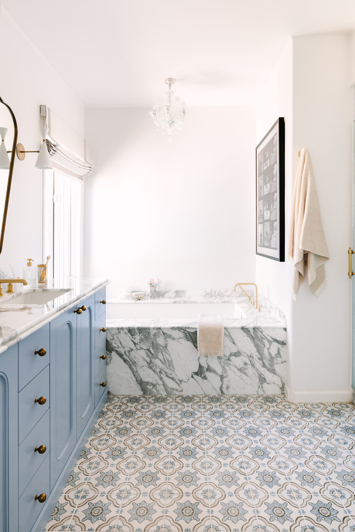 Primary bathroom with a custom vanity in Sherwin Williams Lakeside and rounded edges, Arabescato Corchia marble counters, Italian shield mirrors, Blueprint Lighting Monolith Wall Sconce, patterned floor tile, Murano chandelier, and a built in marble tub.