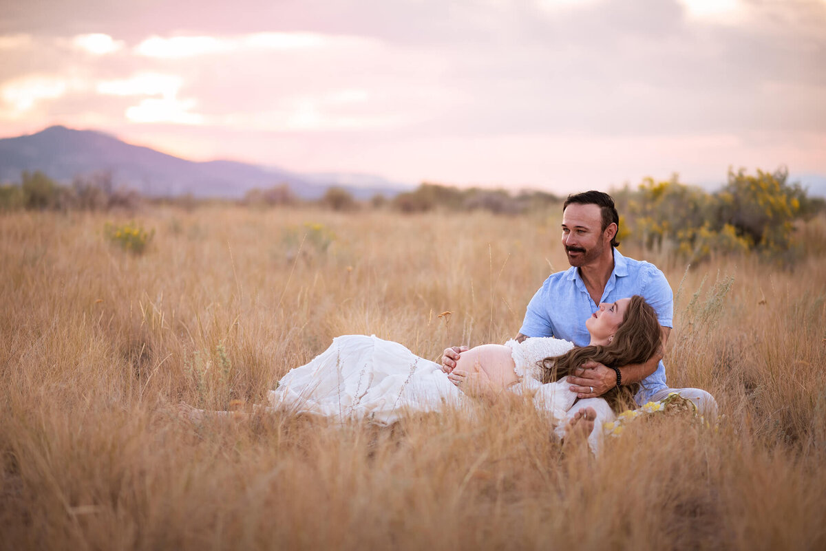 A couple lays in the brown grass in Kamloops during sunset. The man sits while his pregnant wife lays in his lap. They are smiling.