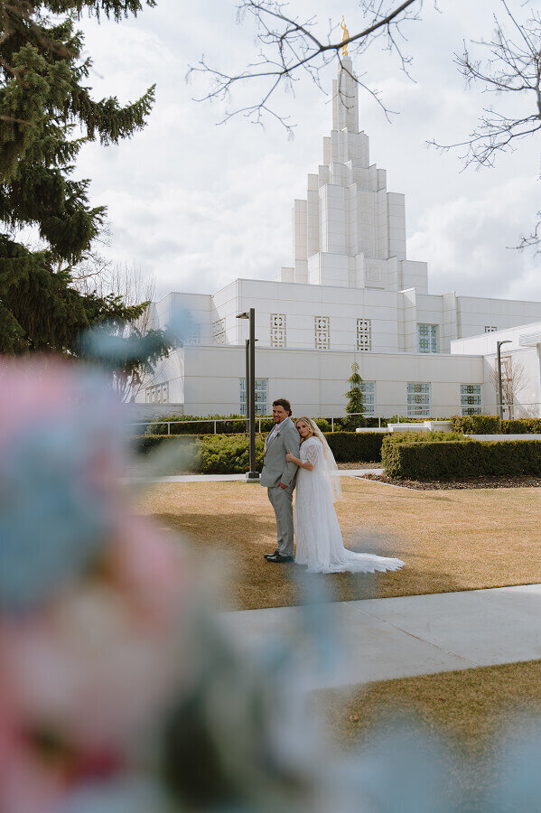 A bride holds the arm of her fiancé as she looks at the camera and smiles. He is facing the same way as she is.