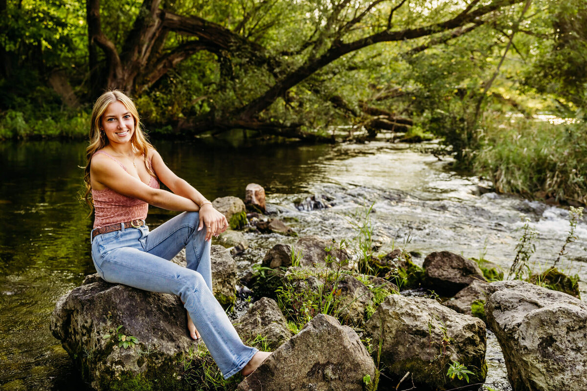 beautiful high school girl in a pink top and jeans sitting on a rock thats in a picturesque creek taken by Ashley Kalbus Photography