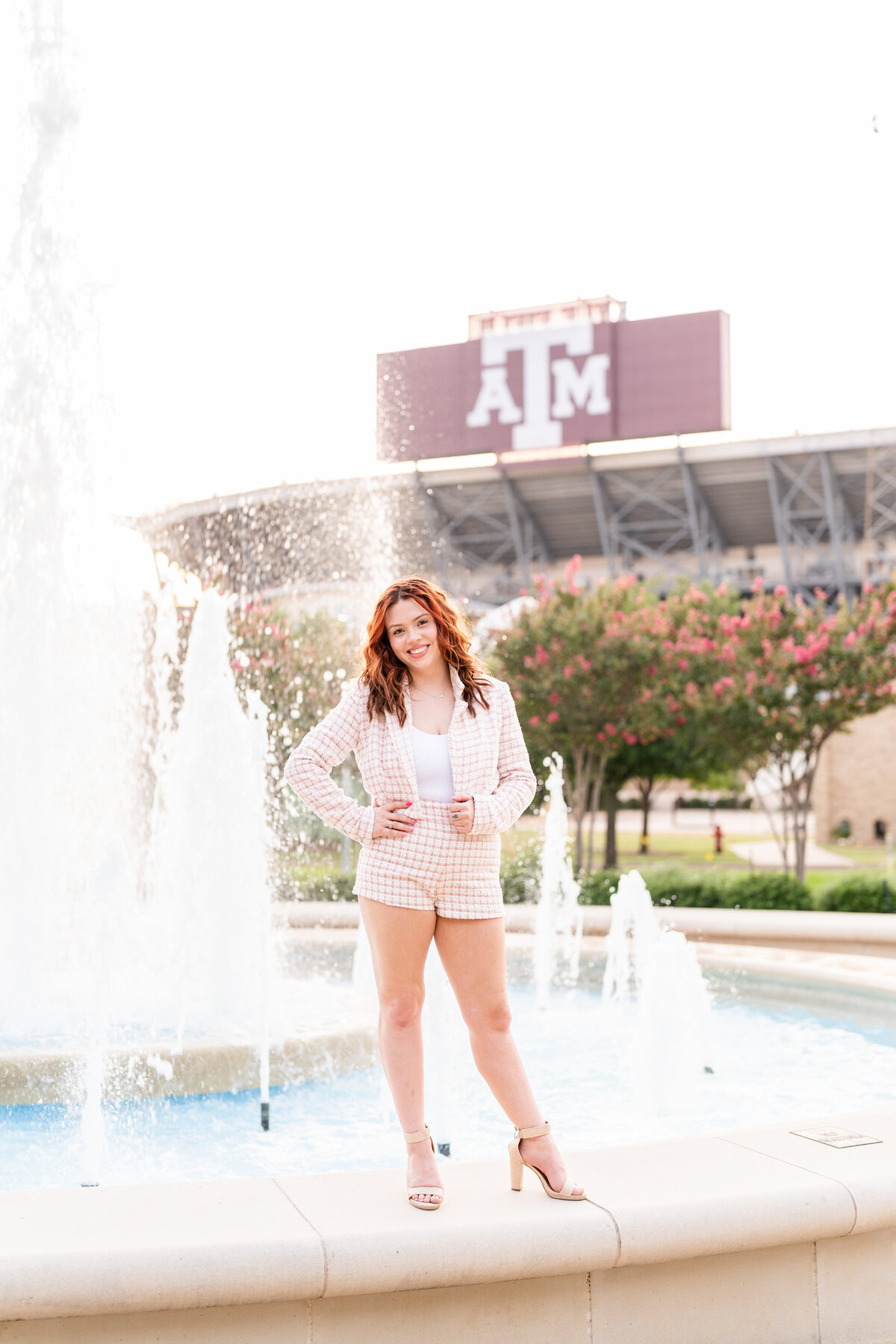 Texas A&M senior girl smiling and wearing cute plaid shorts and blazer with heels and standing on top of fountain edge with Kyle Field in background