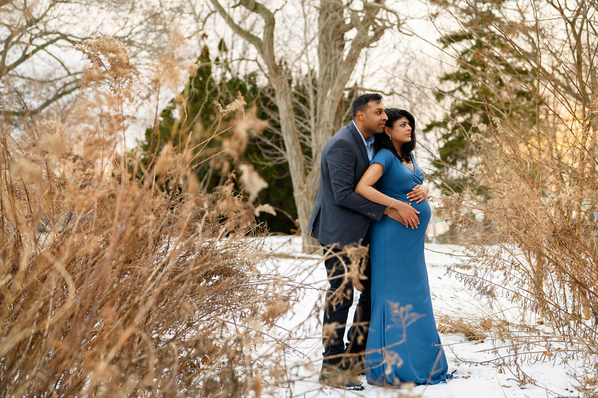 Husband standing behind pregnant wife wearing a blue maternity dress in the snow for her outdoor winter maternity photography session