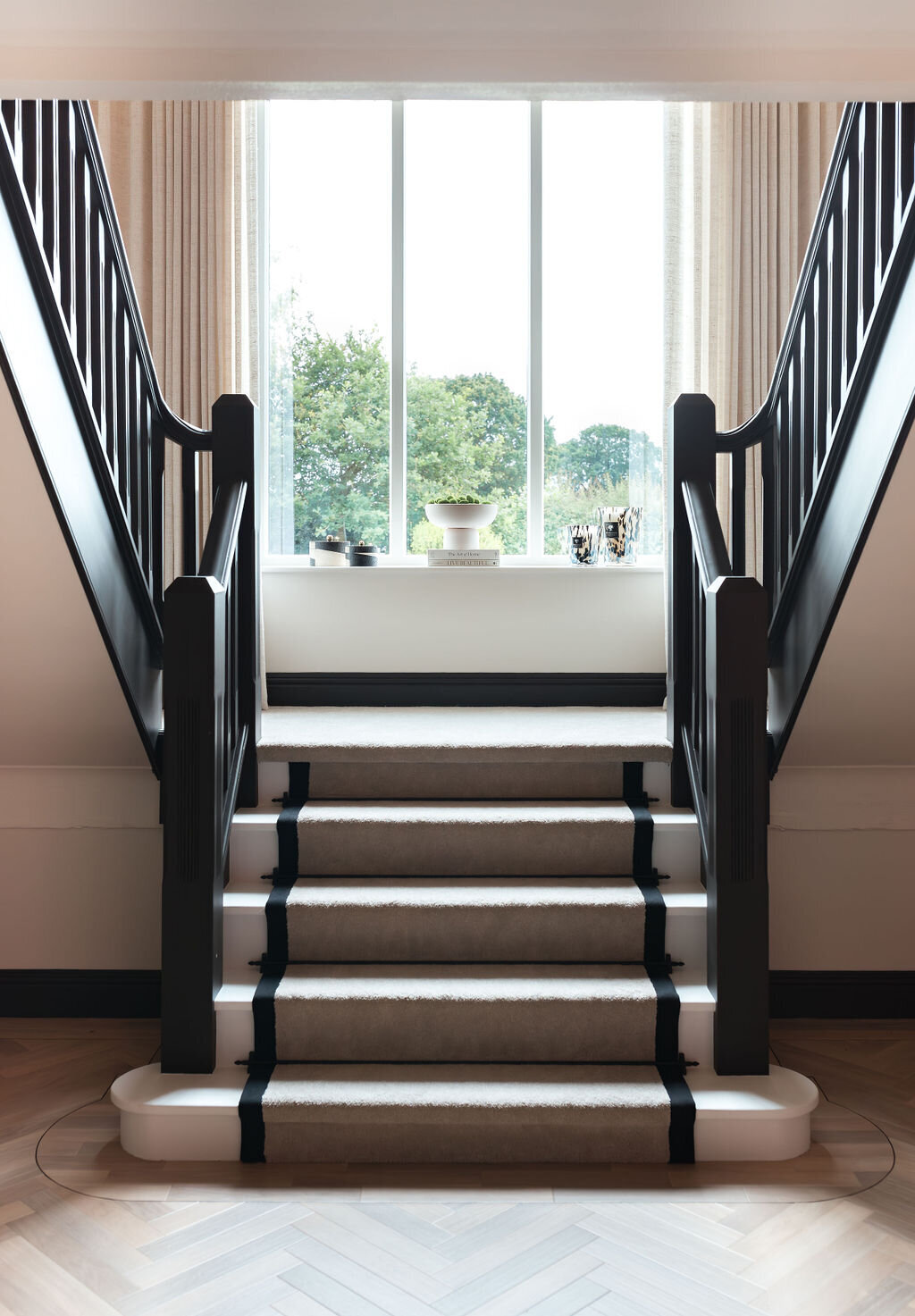 A modern, well-lit white staircase with black railings and beige carpet stair runner with black stripes leads up to a large window. Light-colored curtains frame the window.