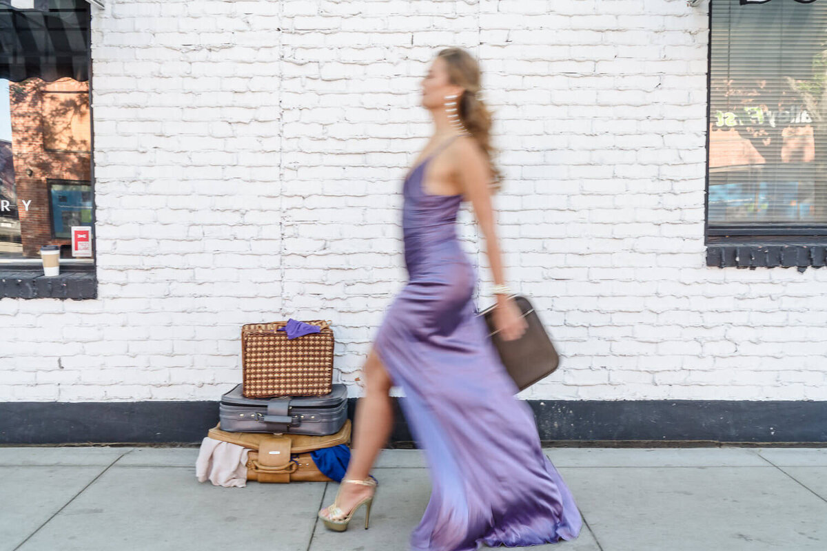 Entrepreneur in flowing lavender dress blurred out walking past white brick wall with vintage suitcase.