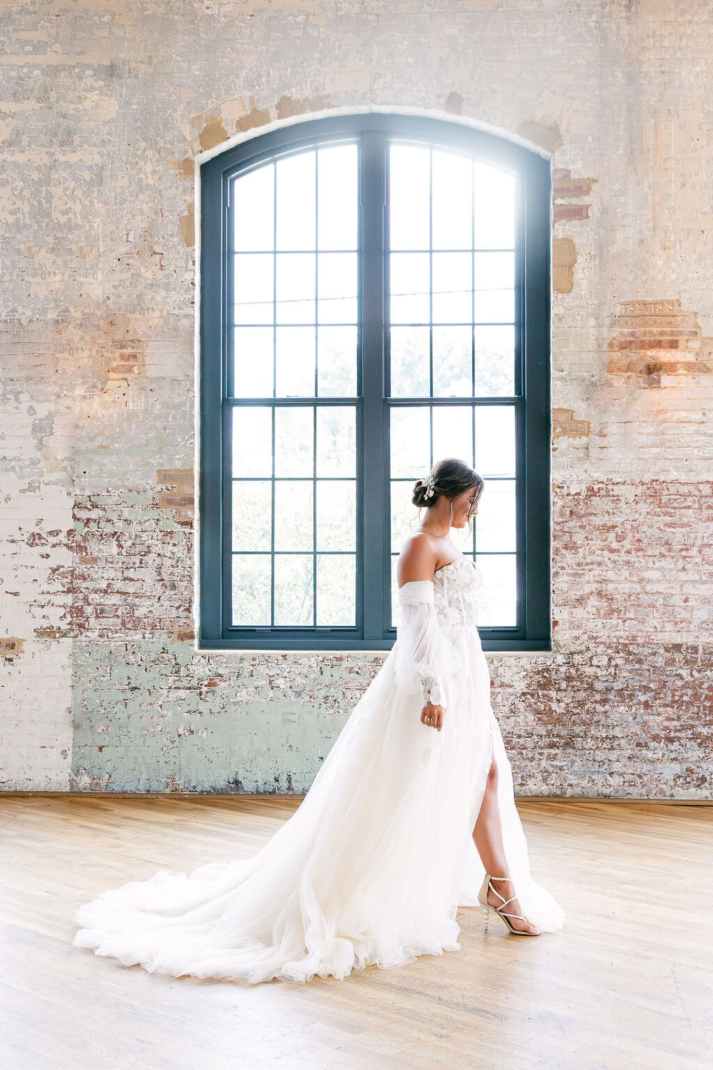 A stunning bride in an ethereal off-the-shoulder gown walks gracefully across an industrial-chic venue in Charleston, SC, with soft natural light illuminating the scene. Wedding photographer Charleston, SC.