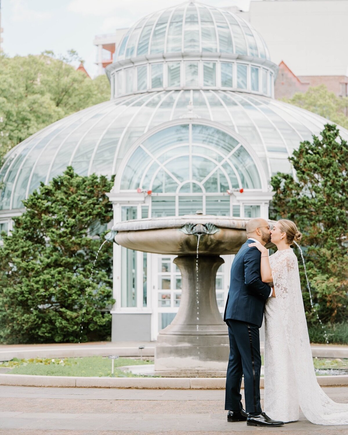 Wedding Photographer RI | A couple shares a kiss in front of a glass conservatory with lush green surroundings and an elegant fountain.