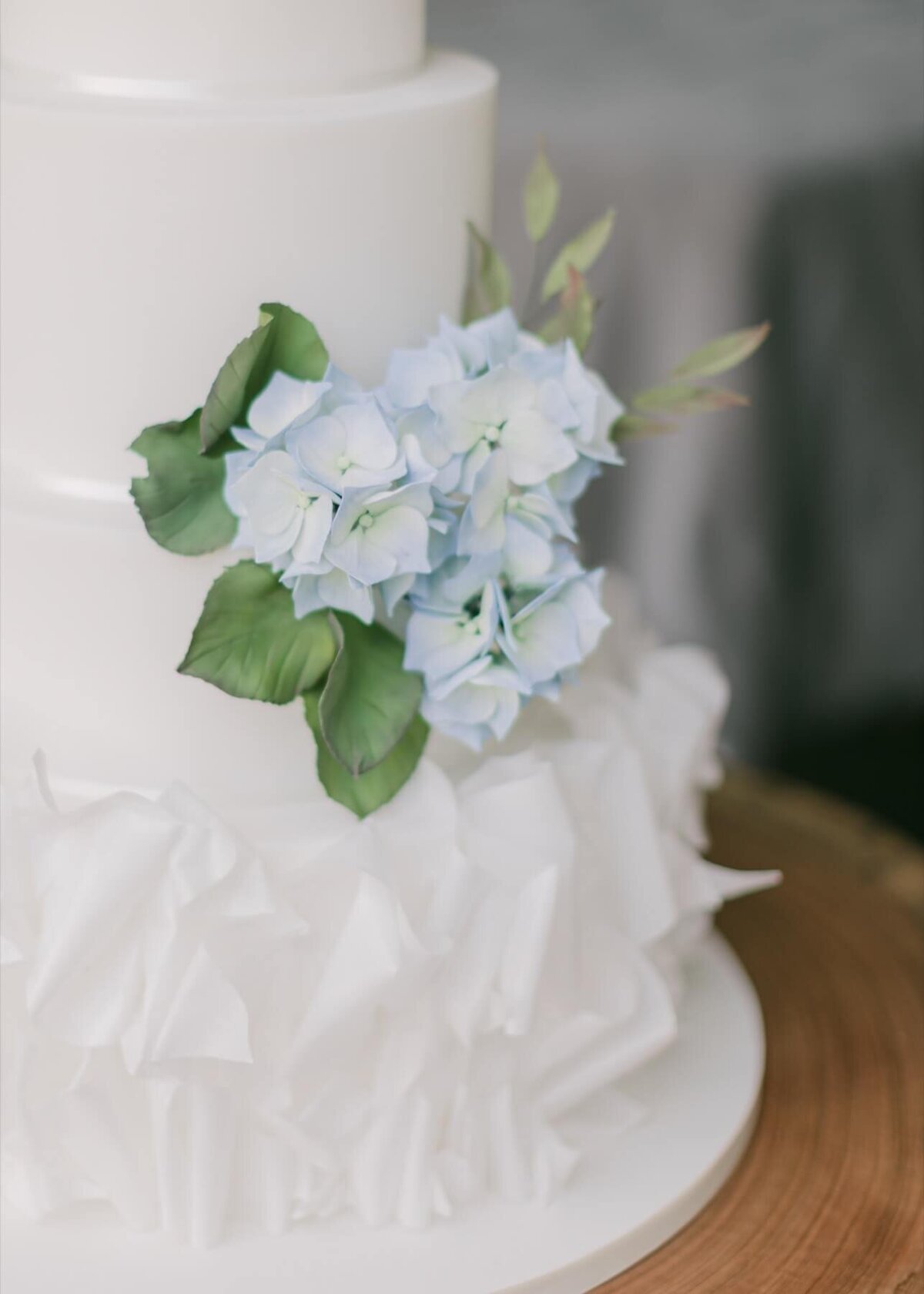 A close up of a white wedding cake showing ruffles at the bottom and blue hydrangea sugar flowers