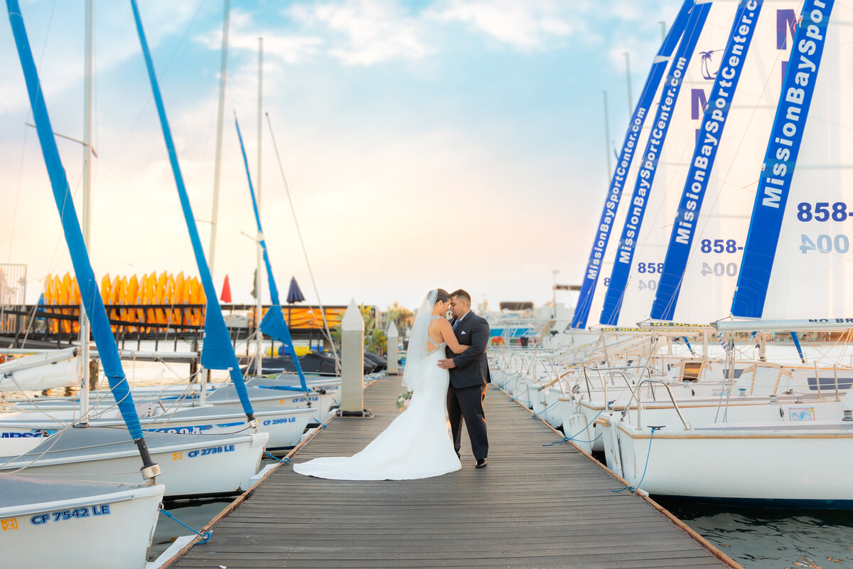 Bride and groom hug each other on a dock with boats on both sides of them in San Diego with stunning sky in the background. Photo by Philippe Studio Pro, wedding photographer.