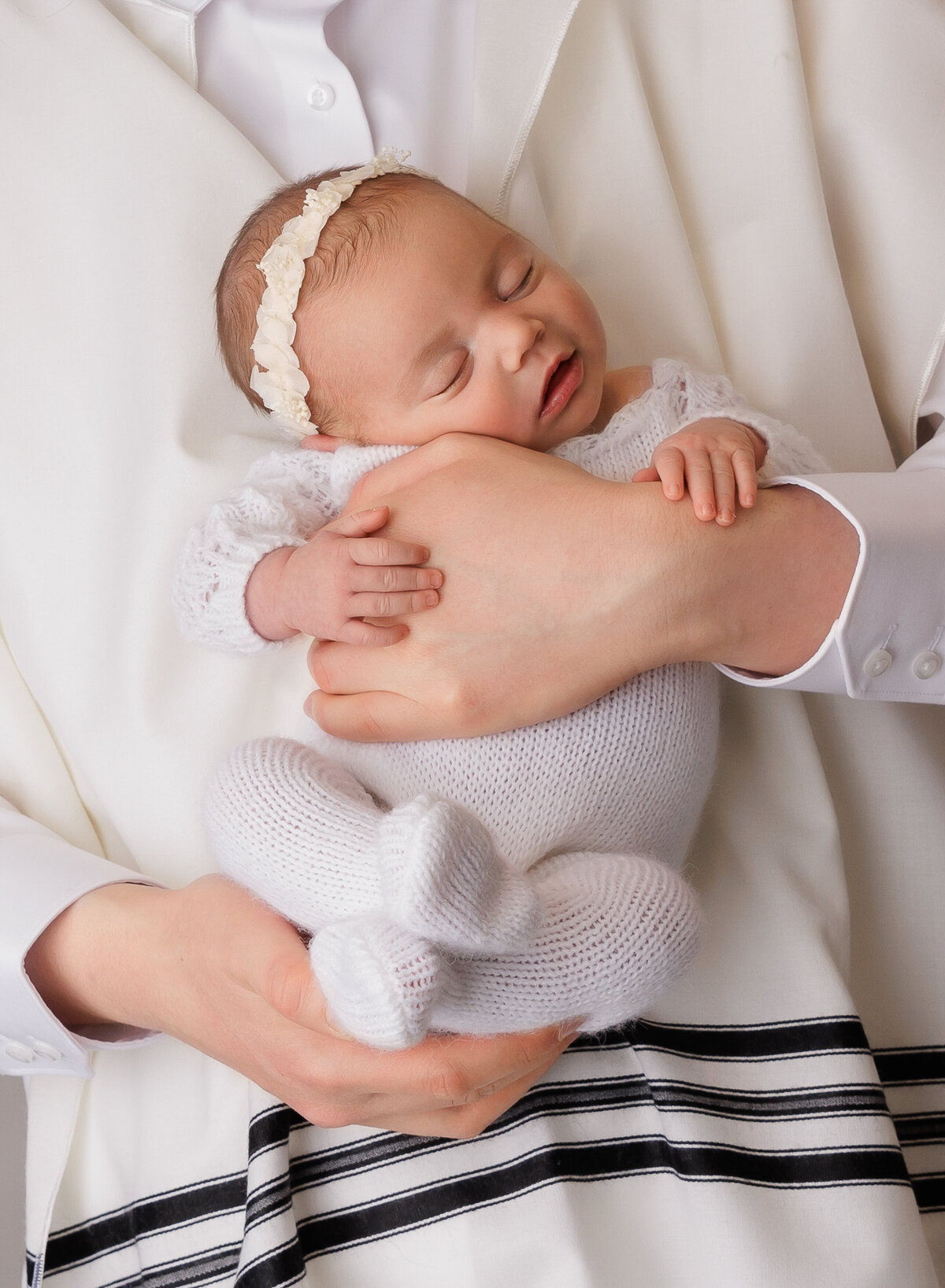 Dad wearing a tallit katan is holding his newborn baby girl against his chest. Baby is facing the camera and her hands are resting atop of dads. Baby is sleeping. Captured by top brooklyn newborn photographer Chaya Bornstein.