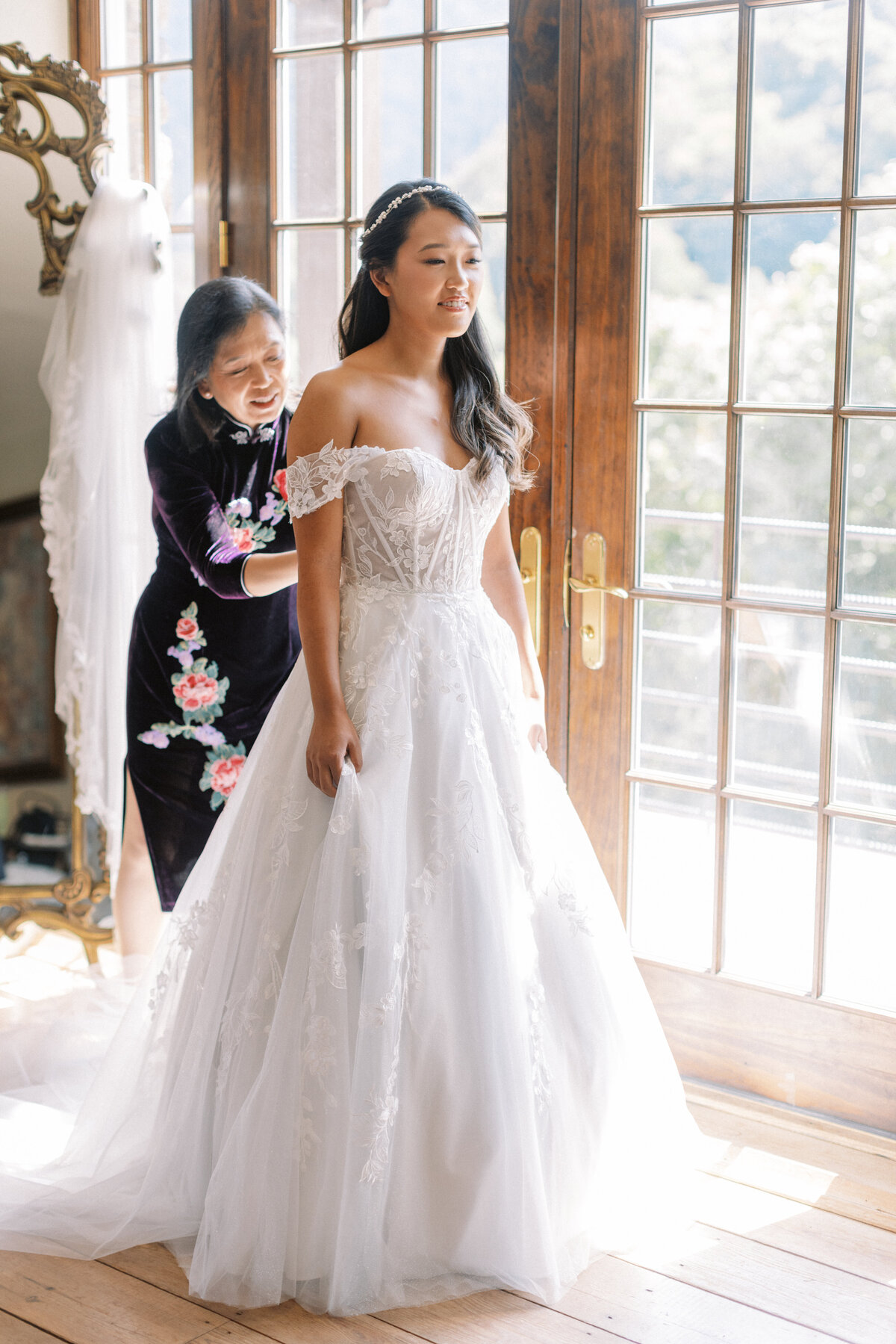 Mother helping bride into her lace wedding gown near tall windows with mountain views at Castle Ladyhawke.