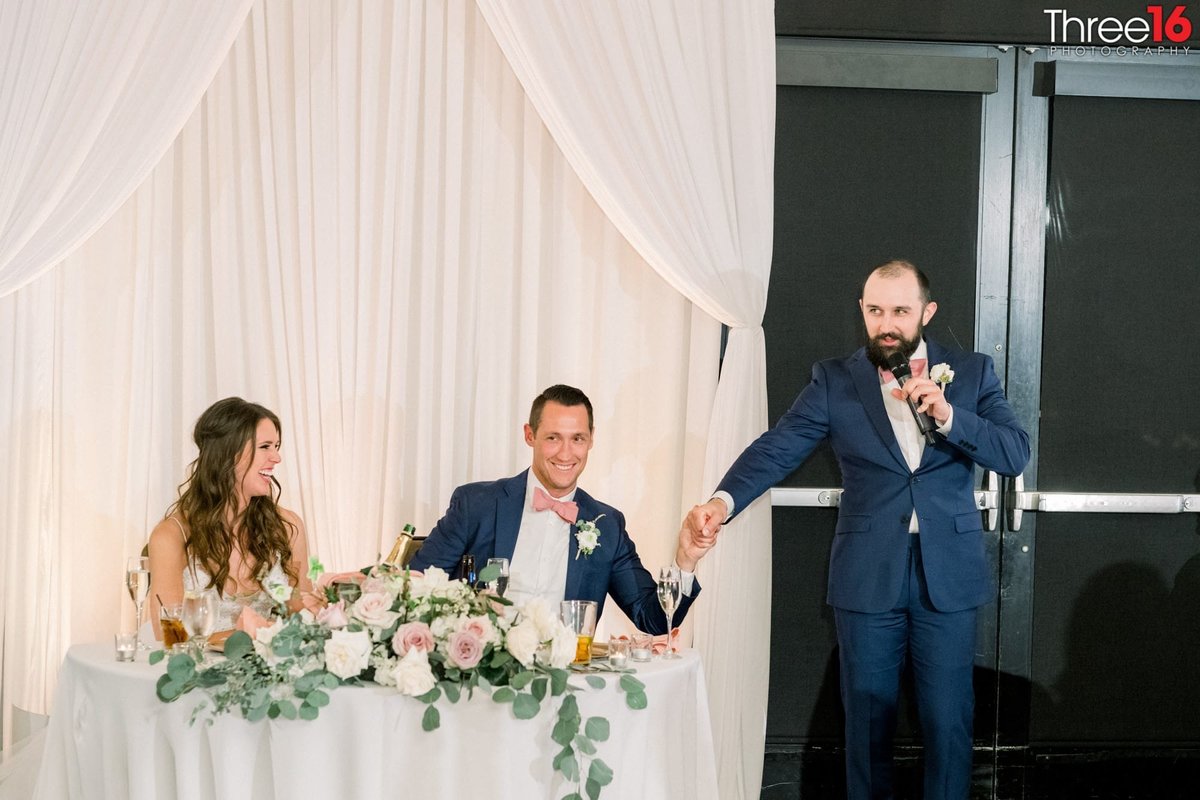 Best Man grabs Groom's hand during the toast as Bride laughs