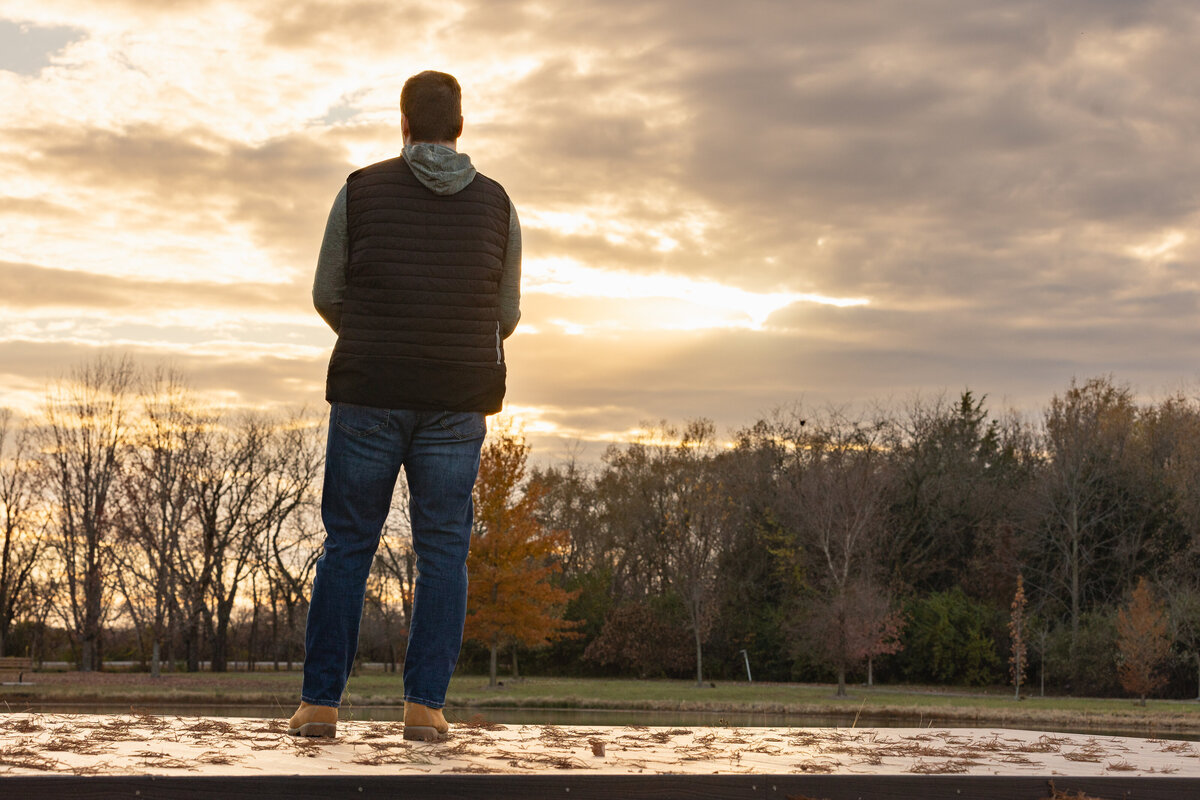 A high school senior stands on a wooden platform, gazing at a dramatic sunset sky with warm golden hues. Dressed in a black vest and jeans, his silhouette against the vast landscape creates a thoughtful and cinematic senior portrait.