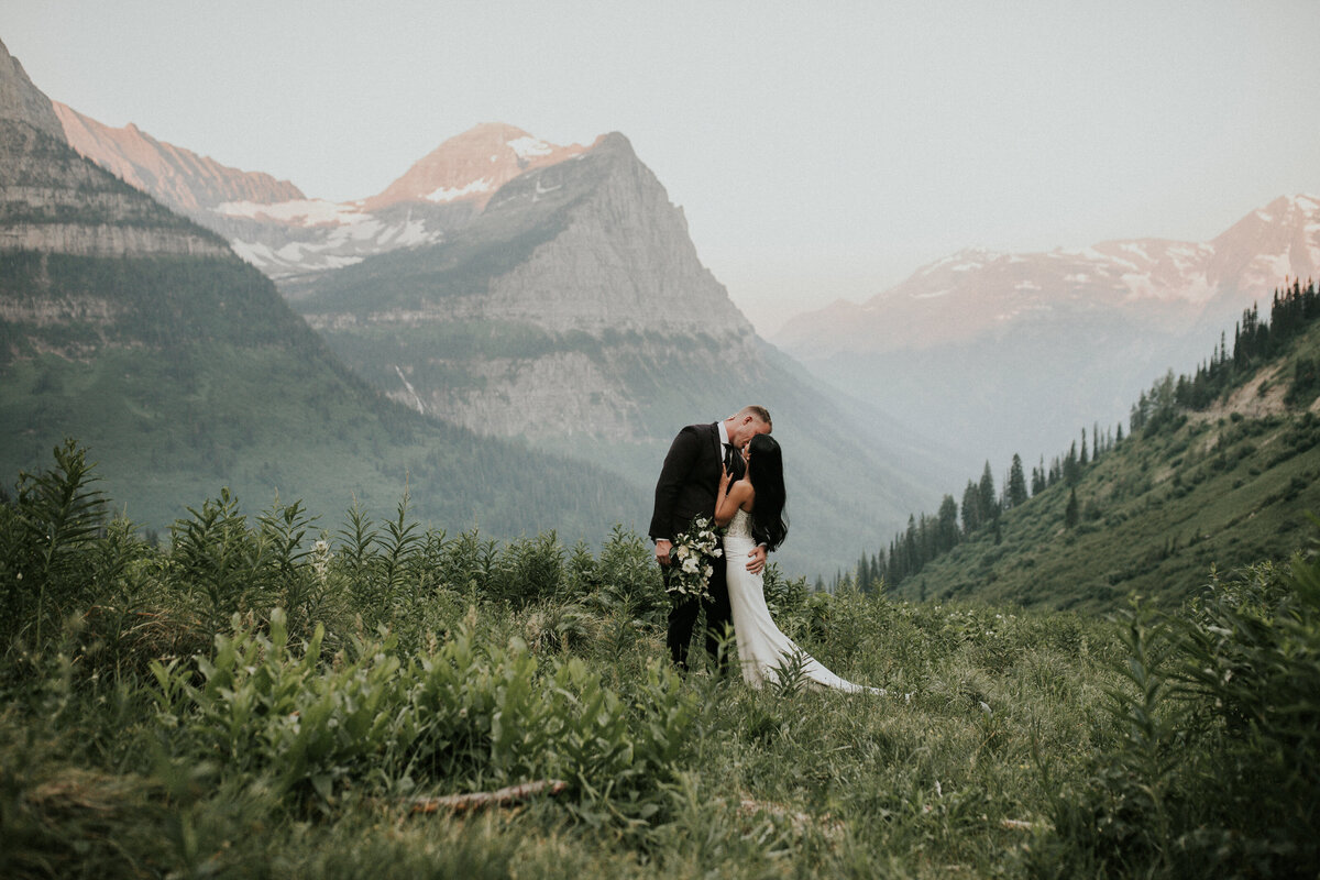 Couple shares a kiss in a grassy tree lined field with a Montana mountain skyscape