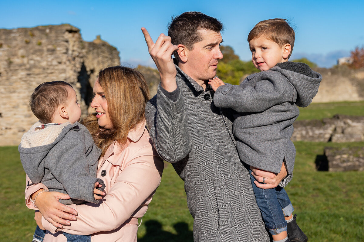 A family of four outdoors at a historical site; parents holding their young sons, dad pointing upwards, on a sunny day.