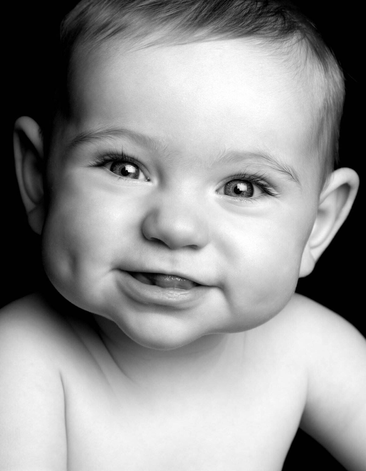 A black-and-white close-up of a smiling baby with chubby cheeks and short hair. The baby has sparkling eyes and dimples, conveying a joyful and innocent mood.