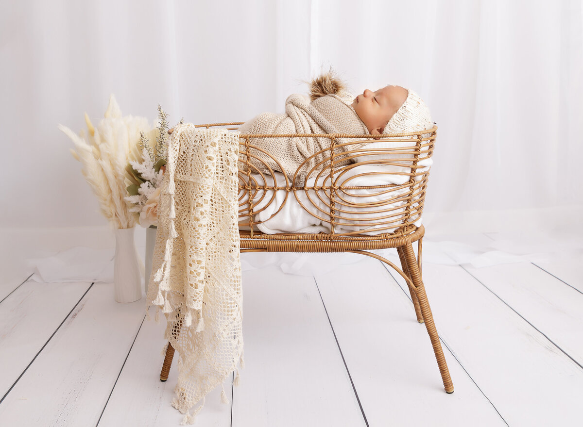 Baby girl is sleeping in a rattan newborn bassinet prop. She is swaddled in taupe with cream fabric draping over the edge. Side profile image of baby. Captured by top Brooklyn newborn photographer Chaya Bornstein