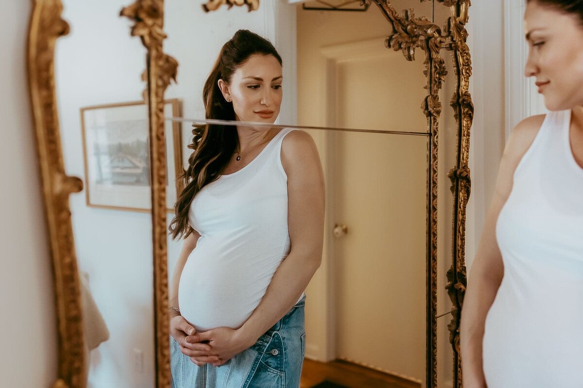Pregnant woman in a white tank top and jeans looks at her reflection in an ornate mirror, standing indoors.