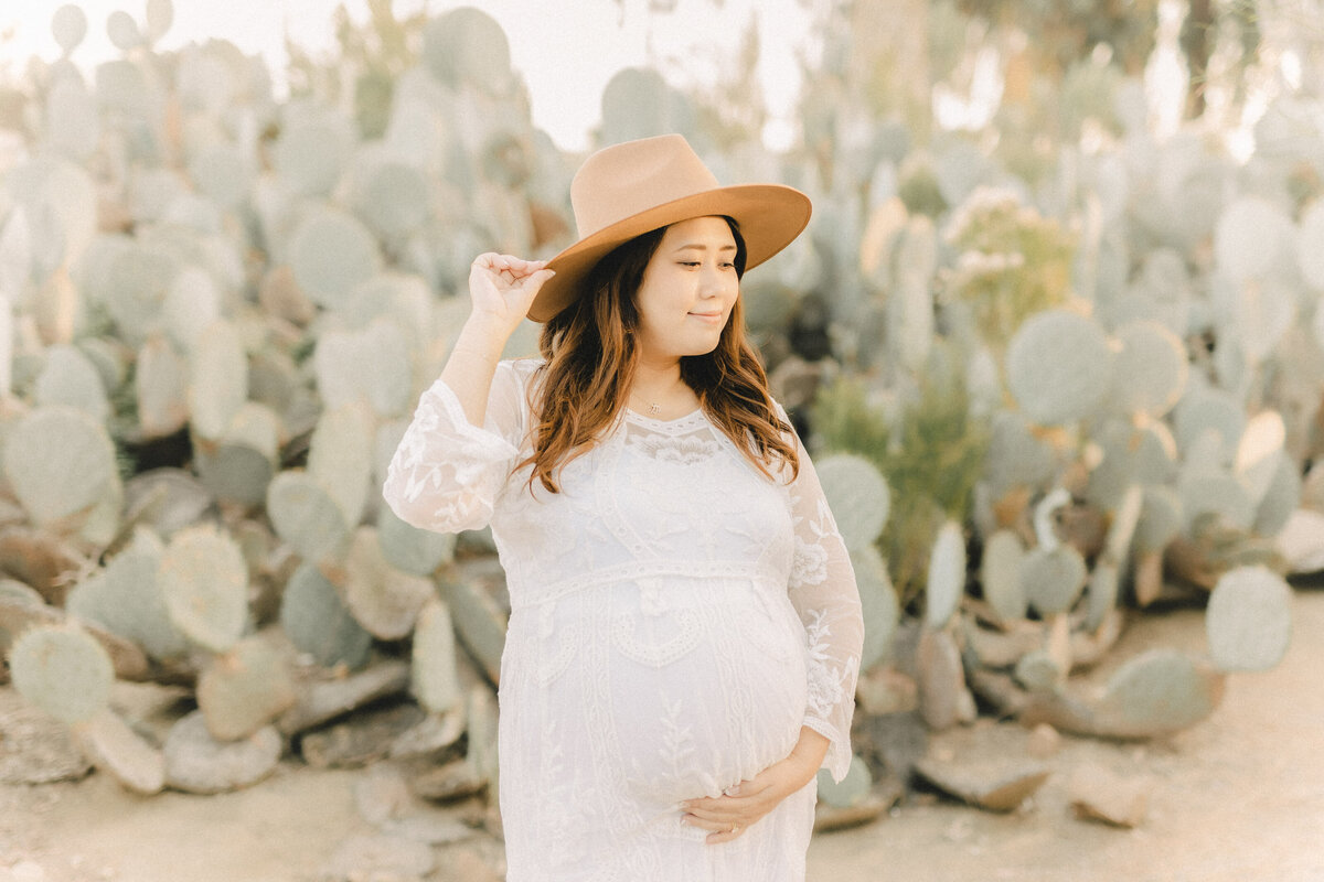 Expecting mother in a white lace gown and tan hat, gently holding her baby bump in a cactus garden during a maternity photoshoot in San Diego.