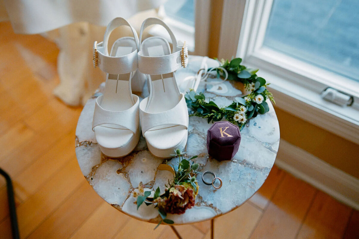 A pair of white high-heeled sandals, floral hair accessories, two rings, and a purple ring box with a gold K sit on a marble-topped table by a sunlit window—styled perfectly for a film photographer NJ to capture timeless details.