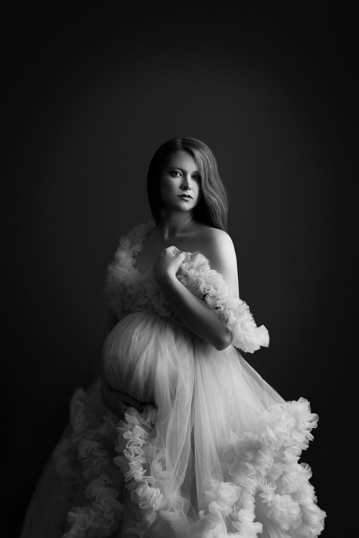 Black and white maternity photo taken by Katie Marshall, recognized as the best Main Line maternity photographer. The image features a woman in an elegant tulle maternity photoshoot gown with embellished ruching at the hem, gracefully draped off her shoulders. She delicately touches her shoulder with one hand, while the other cradles her baby bump. With her hair pushed behind her back, she gazes over her shoulder towards the camera, creating a timeless and evocative maternity portrait.