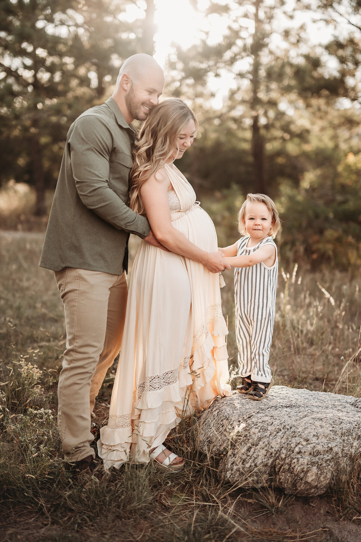 little boy standing on a rock holding his mom's hands while dad hugs her back