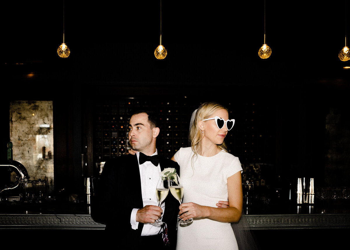 Trendy direct flash portrait of bride and groom during cocktail hour, bride wearing heart shaped sunglasses captured by TkShotz, modern wedding photographer and videographer in Calgary, Alberta. Featured on the Bronte Bride Vendor Guide.