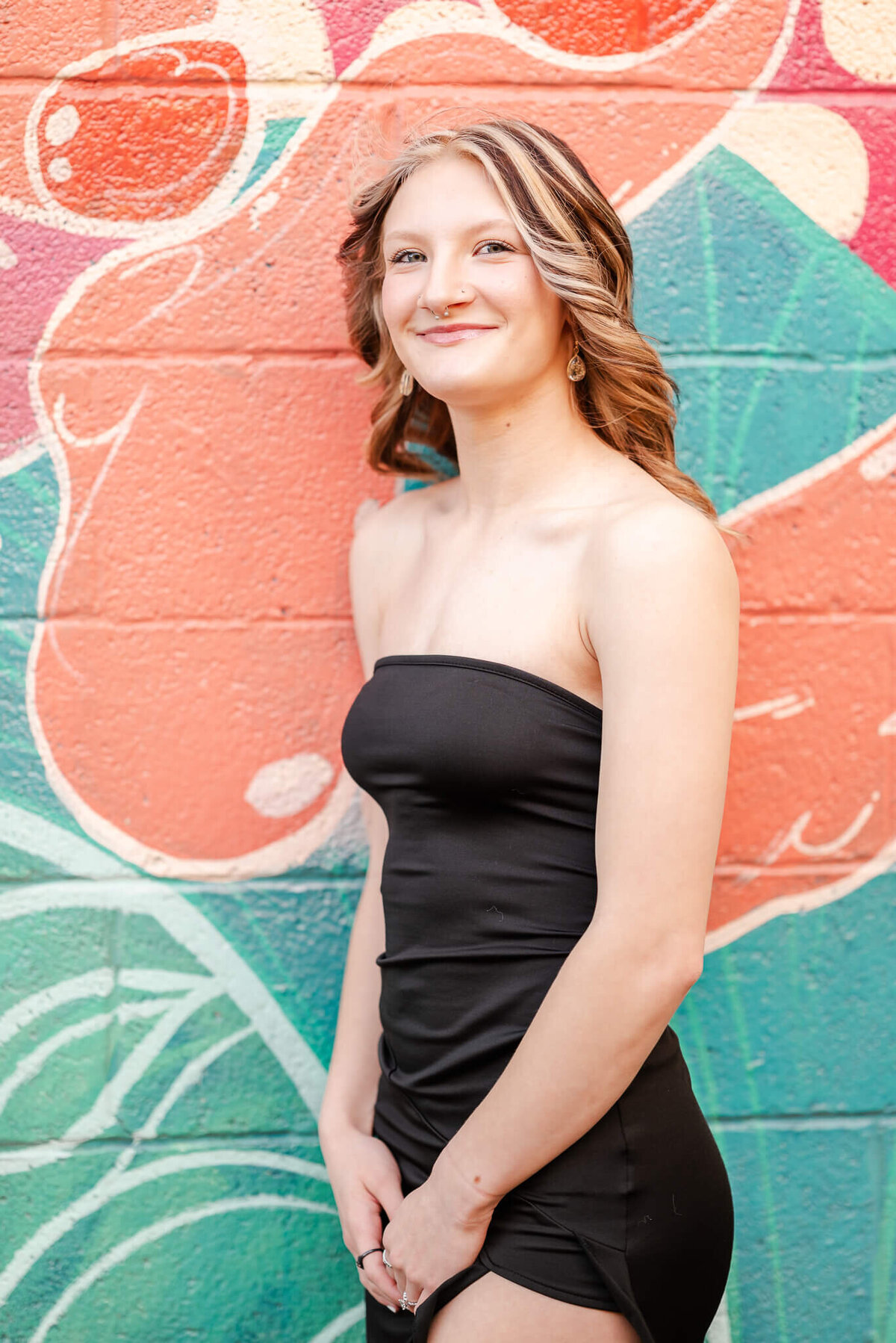 A high school senior, wearing a black dress, leans against a wall with a colorful mural. She is having her senior session with Chesapeake Senior Photographer, Justine Renee Photography.