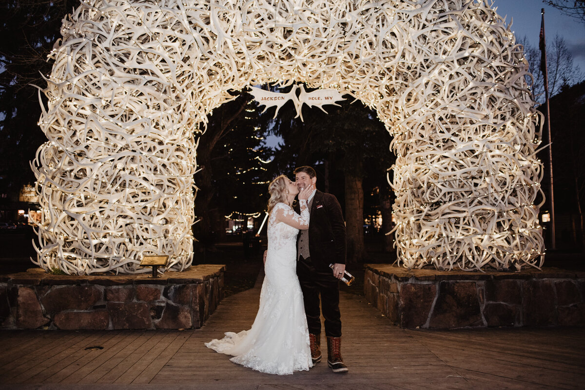jackson hole wedding with bride and groom kissing under the deer antler arch in downtown at night with the bride kissing her groom on the cheek as he laughs