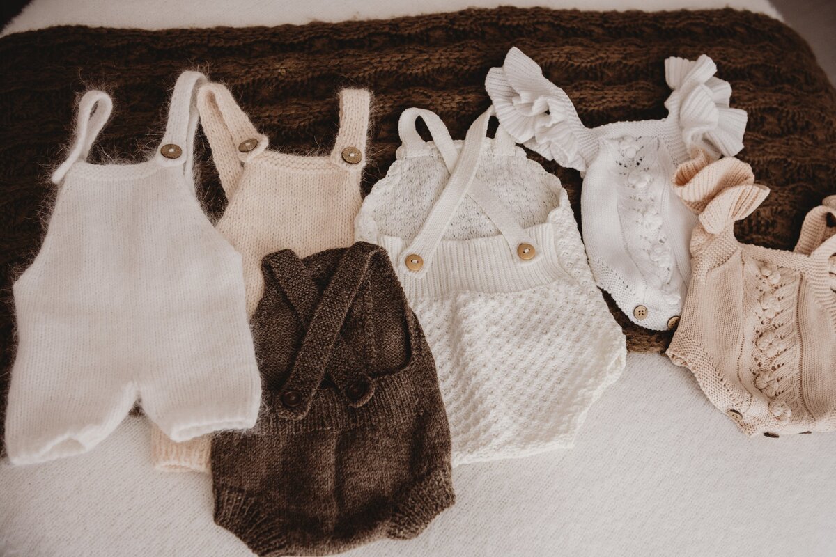 A selection of knitted baby rompers, perfect for your South Coast newborn, in various neutral colors including white, beige, and brown, laid out on a soft, textured surface.