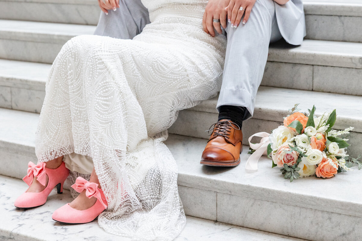 A close-up of the couple sitting on the marble steps, showcasing the bride’s delicate lace gown and pink heels, with her bouquet beside them.