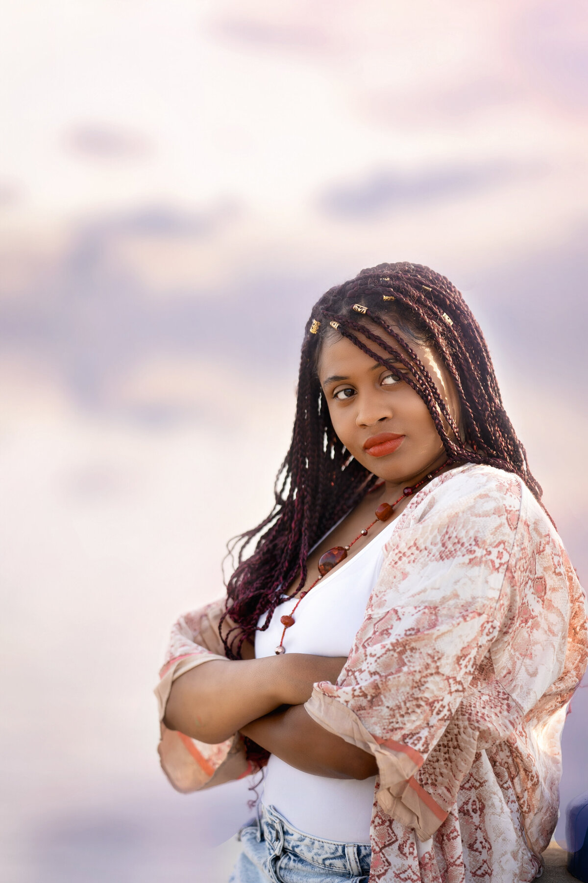 Black high school senior girl wearing a white tank top and jean shorts.  She has a floral kimono on.  She has long braids and red lipstick.  Her arms are folded across her chest and she is looking off to the side.  The sky is blurry behind her.