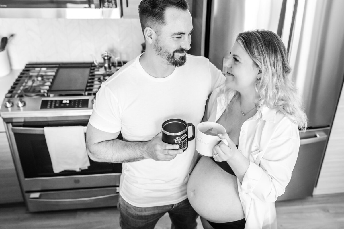 A black-and-white photo of a couple standing in the kitchen, each holding a coffee mug, and gazing at each other with smiles; the woman is pregnant and wearing a white shirt.