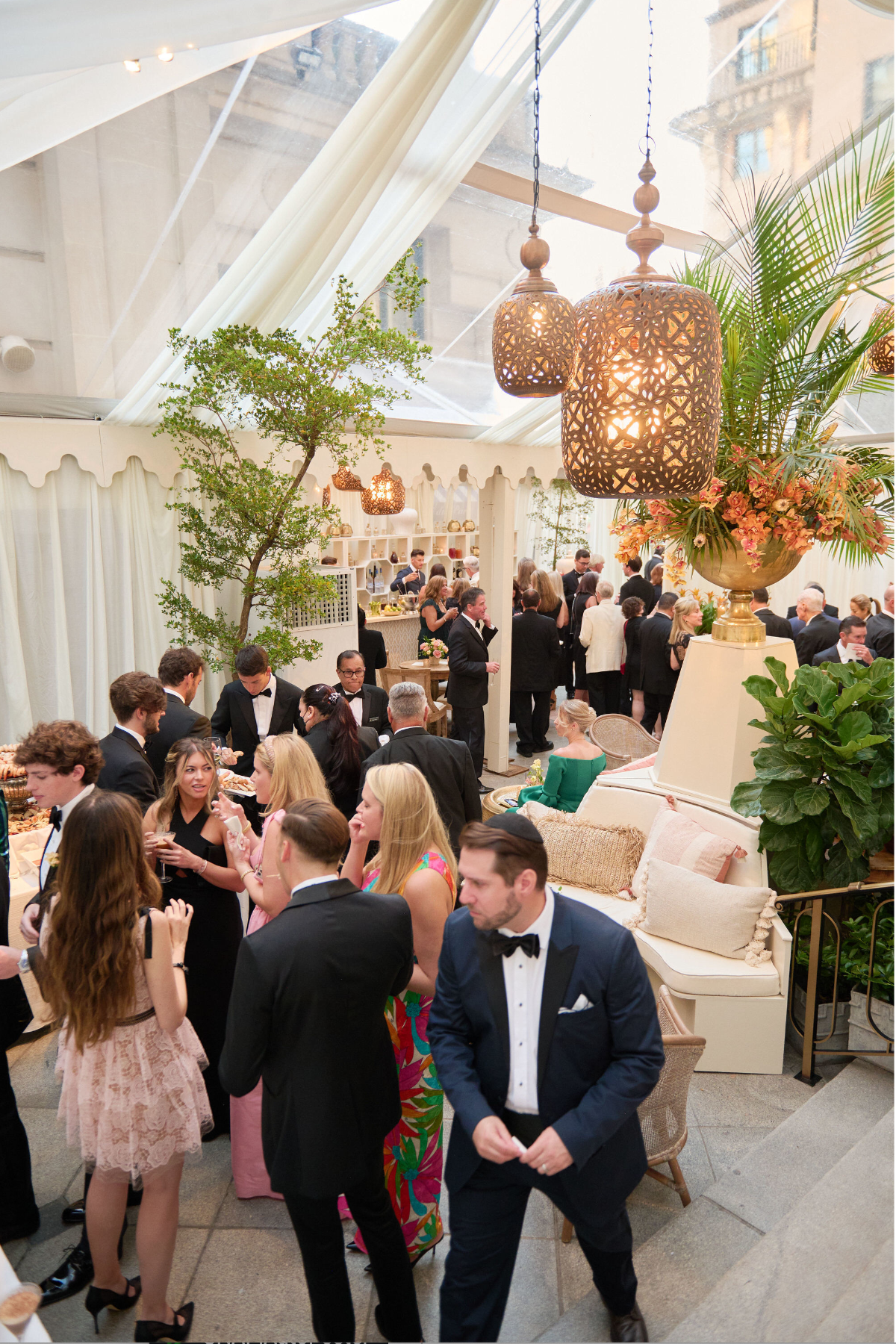A lively formal gathering takes place under a transparent tent with hanging lanterns and lush greenery. People in elegant attire, including suits and evening gowns, engage in conversation, laughing and enjoying the sophisticated atmosphere.