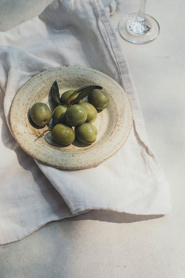 Assiette en céramique avec des olives vertes posée sur une nappe en lin blanc.