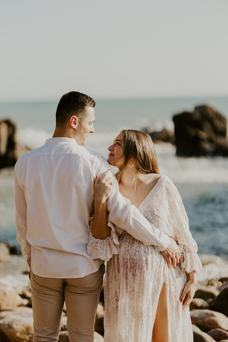 Couple enlacé face à la mer et aux rochers. La femme enceinte, dans une robe en dentelle blanche regarde son mari dans les yeux et lui tient le bras. Photo prise par Laura, photographe grossesse en Vendée.