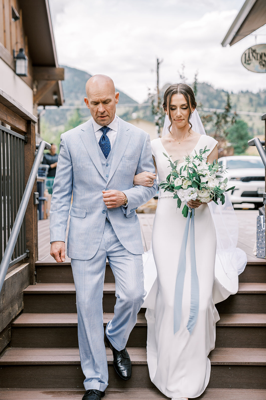 Bride walks down the aisle with her father carrying a white and greenery bouquet with light blue accents.