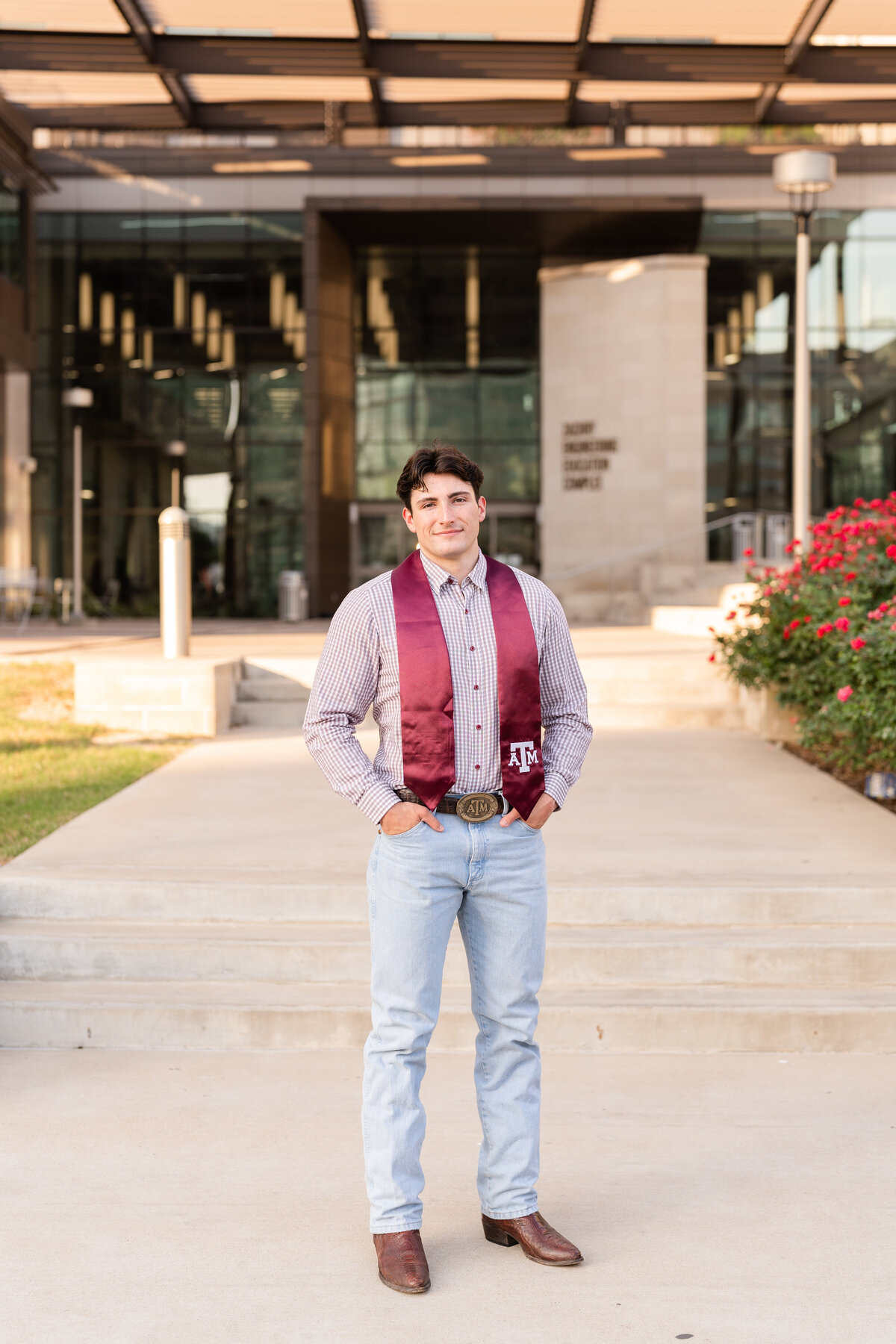 Texas A&M senior guy standing with hands in pocket and light wash jeans with button up top and Aggie stole in front of Zachry Engineering Complex