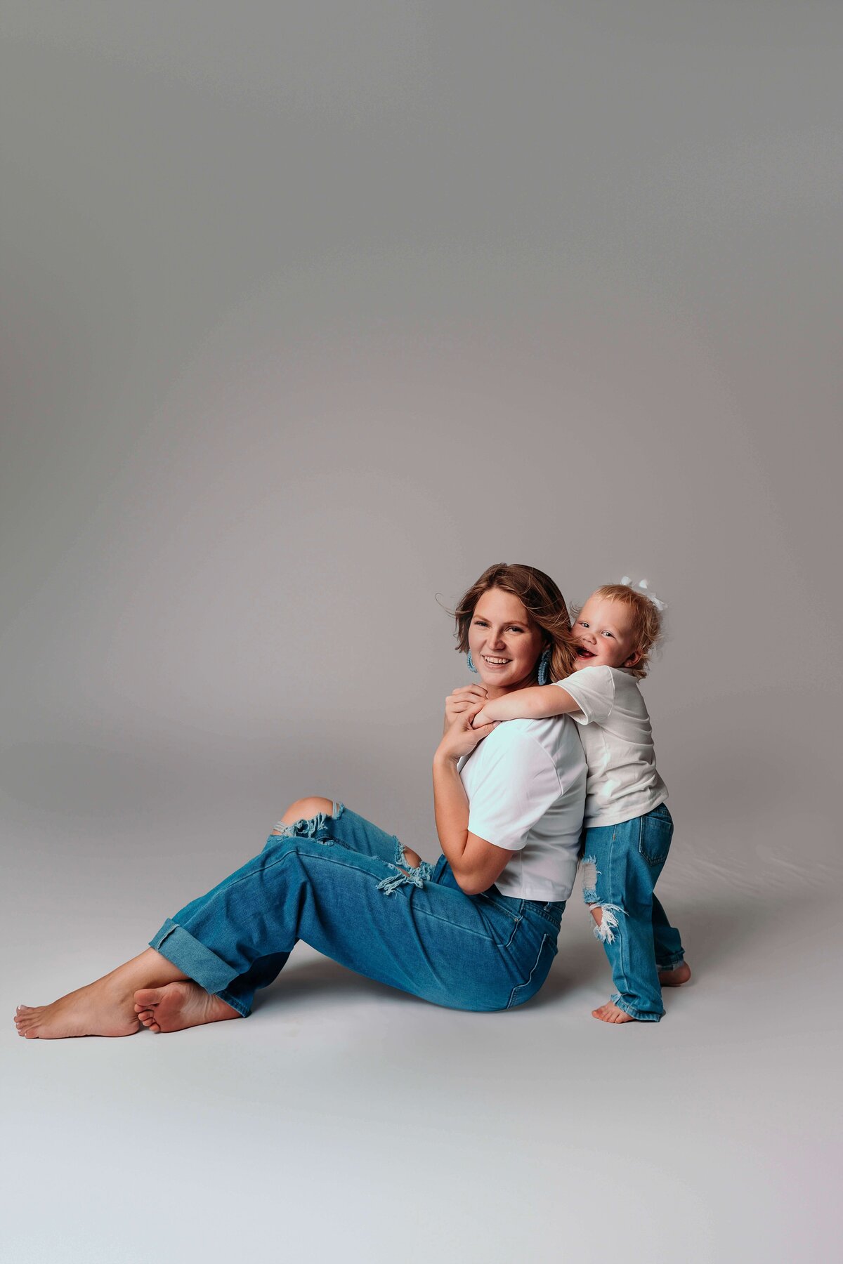 A young mom and her toddler daughter pose for a playful portrait by Mobile, AL Motherhood Photographer. Both are wearing matching jeans and shirts, and are barefoot. The child stands behind her seated mom, leaning against her back for a quick embrace, while the mom looks at the camera with a loving expression