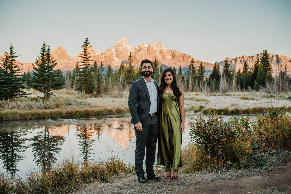 photographers in jackson hole photographs Grand Teton engagement pictures with couple in front of a creek an engaged couple wearing formal attire pose in the Tetons for their formal fall engagement session