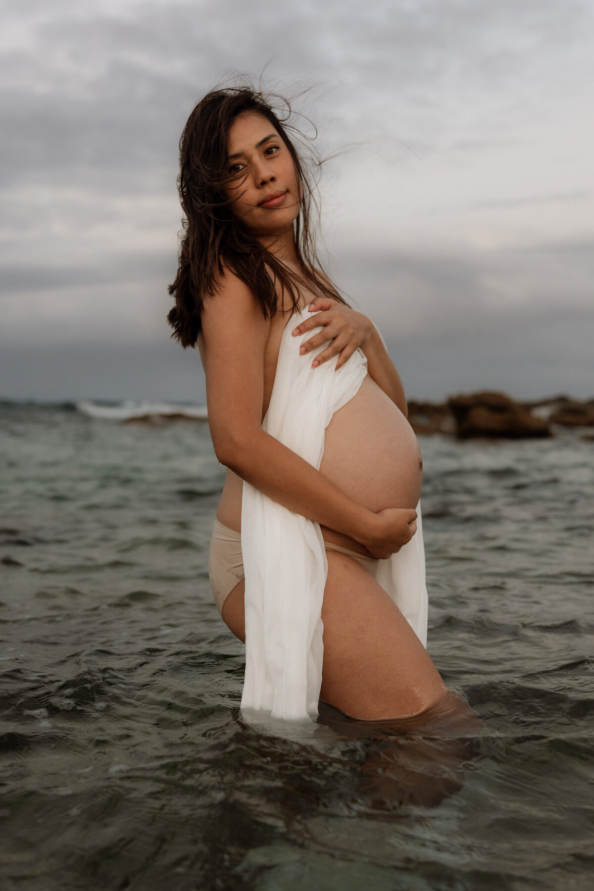 Pregnant woman standing in shallow ocean water during a maternity beach session in Sydney, wrapped in a white cloth and looking confidently at the camera.