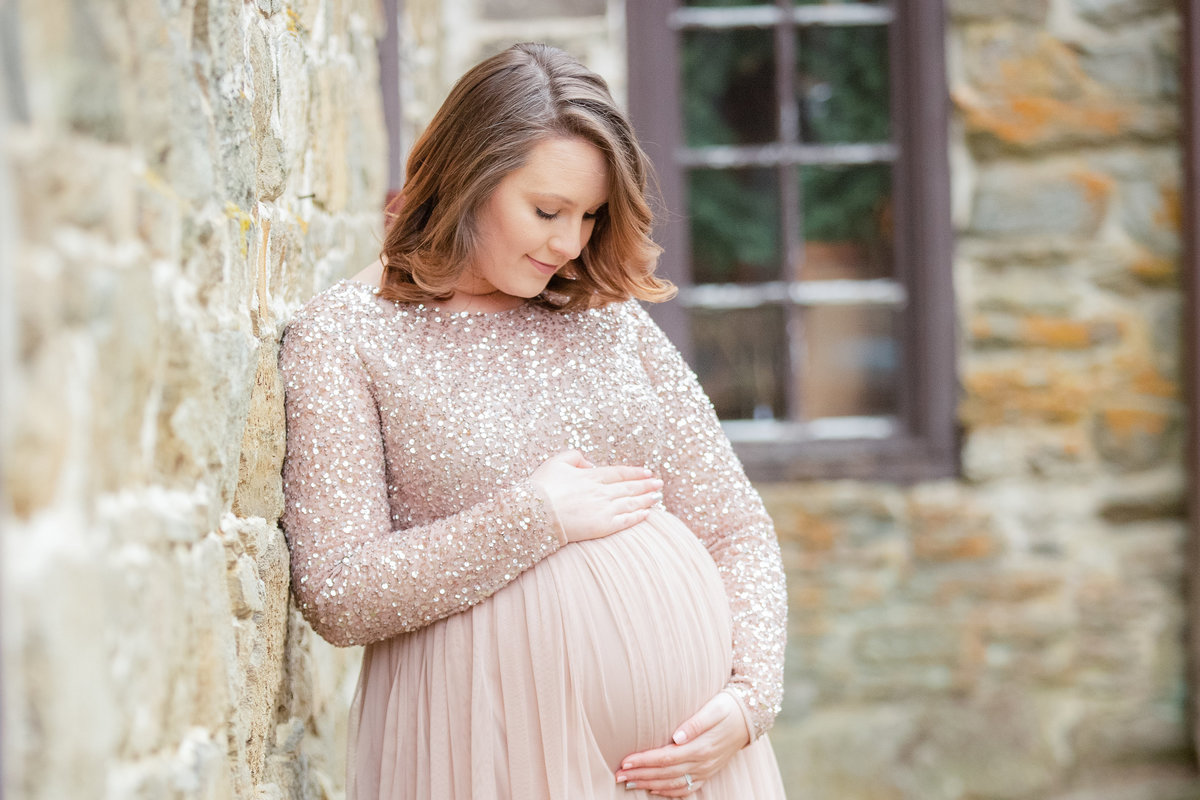 Beautiful pregnant woman in pink gown looks down lovingly as she embraces her pregnant belly during maternity photo shoot