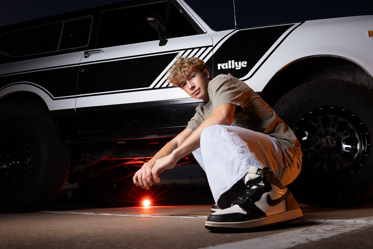 A senior crouches beside his vintage truck, showcasing his unique style in senior photos Dallas.