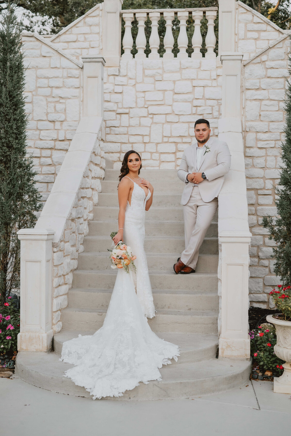 Bride in elegant lace wedding gown holding bouquet and groom in light suit posing on stone staircase at luxury wedding venue, photographed by Kyrsten Ashlay Photography