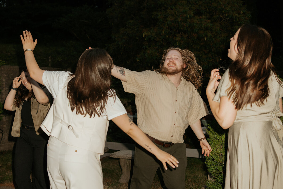 Wedding guests dancing and having fun outdoors at a reception, capturing candid moments of joy and celebration. The group, including a man with long curly hair and a beige shirt, laughs and moves energetically under the night sky, reflecting the lively and festive atmosphere of the event.