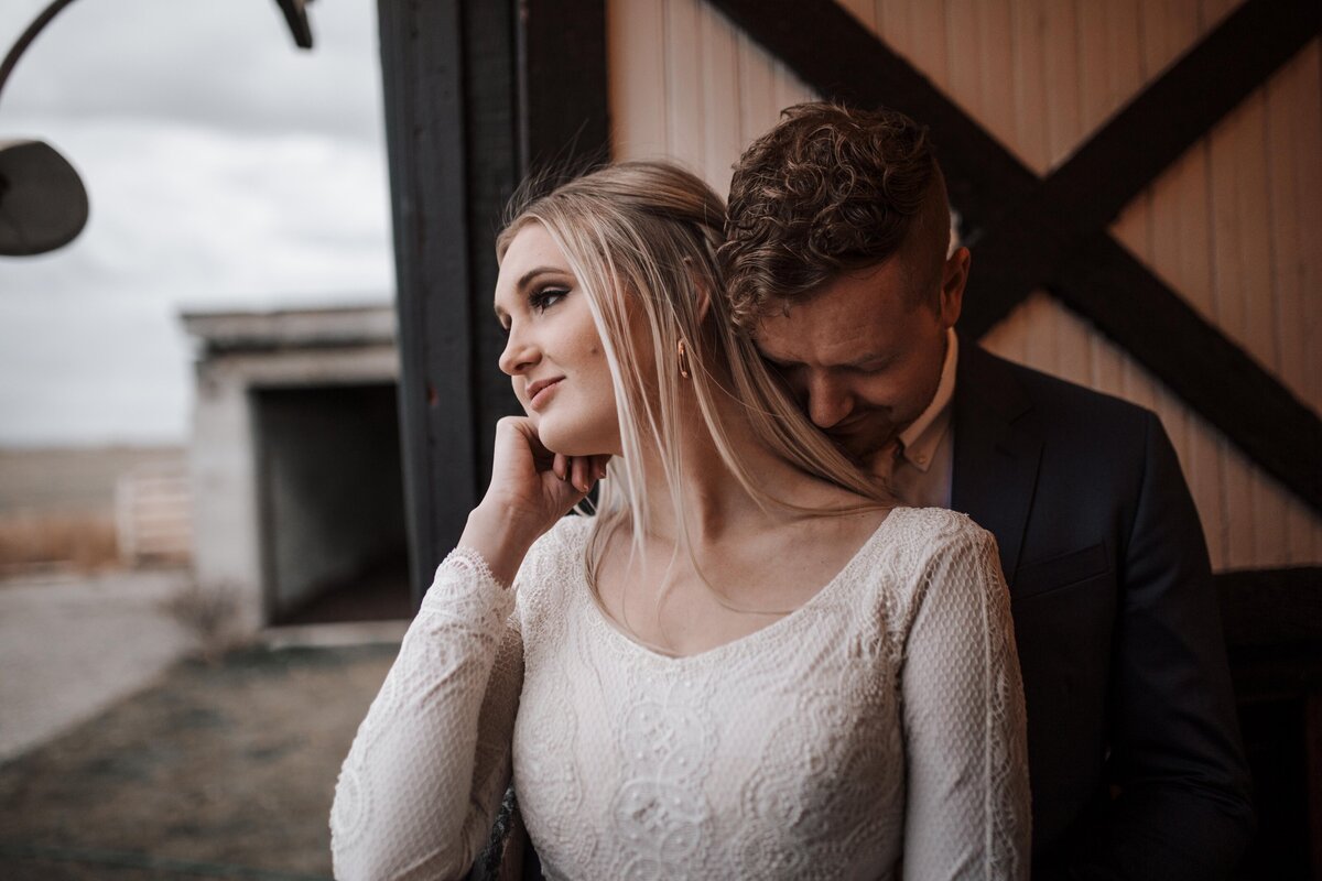 Idaho Falls wedding photographer documents Idaho barn wedding with bride looking out on a pasture from a barn as her groom stands behind her and kisses her neck