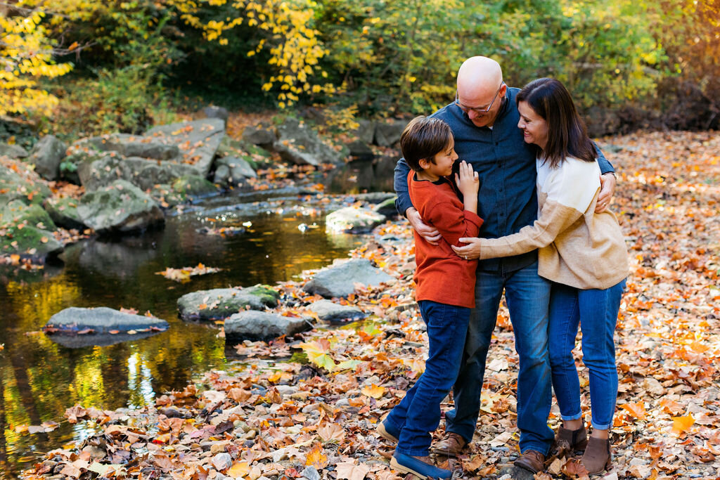 A family of three hugs next to a pond.