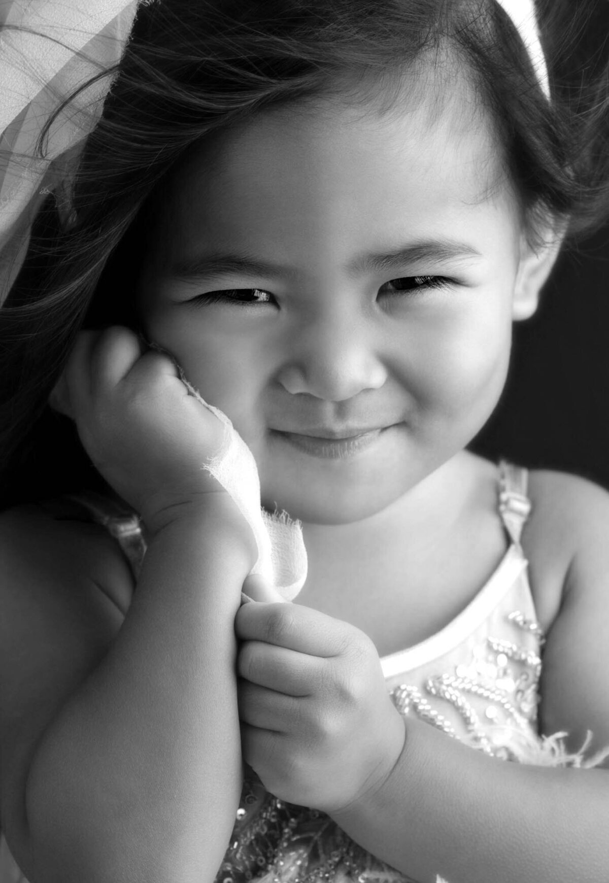 Smiling child in black and white, holding a soft cloth to their cheek. Wearing a detailed top, they exude innocence and joy against a dark backdrop.