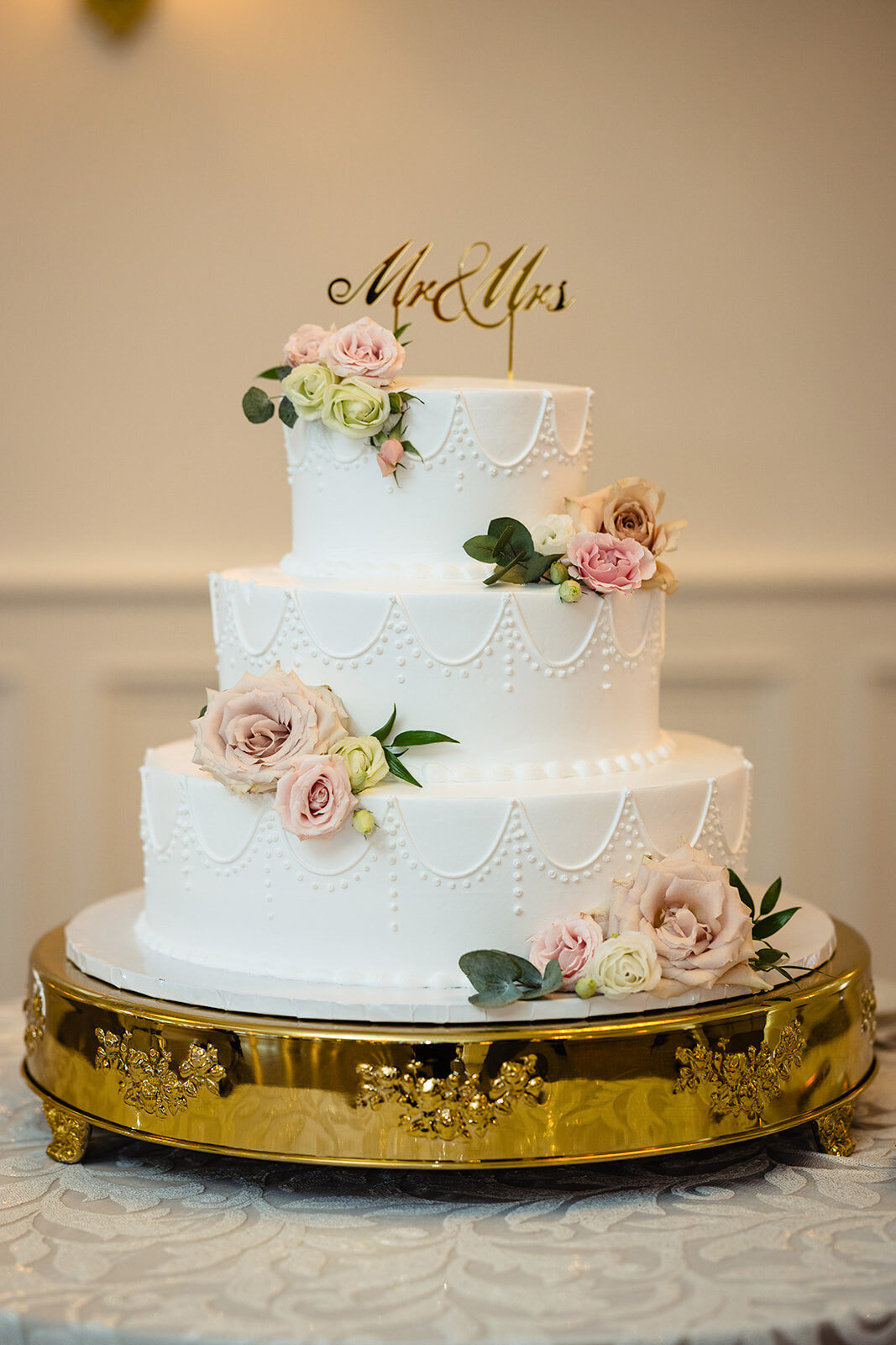 A white three-tiered wedding cake with 'Mr & Mrs' topper and adorned with delicate flowers on a gold stand