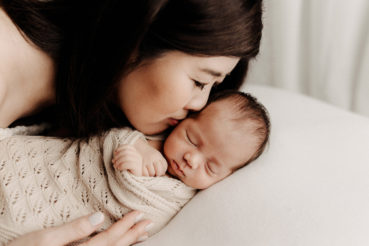 Mother gently kissing her swaddled newborn’s head, both resting on a cream blanket, capturing a tender and intimate moment.
