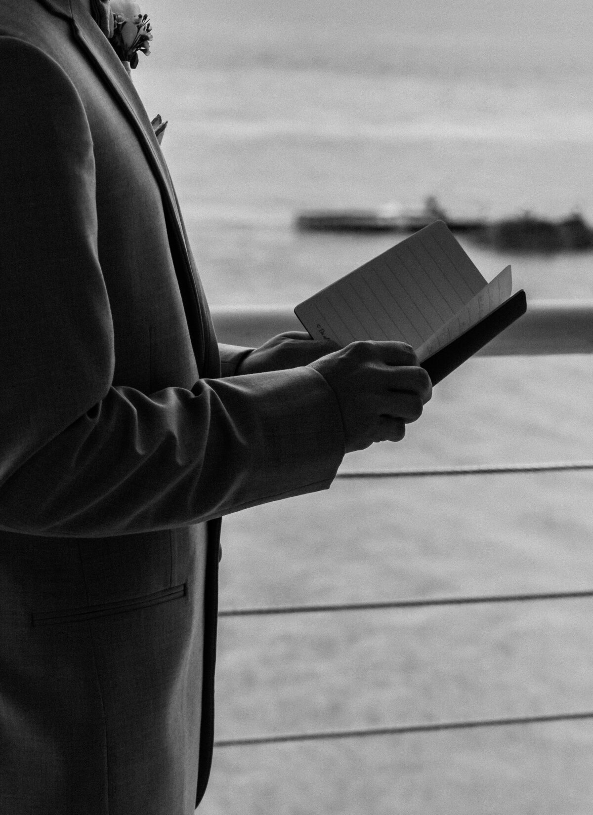 A close-up, black and white shot of a groom in his suit reading his vows, with the waterfront background visible from the Lesner Inn in Virginia Beach, captured by Quinn Photography LLC.